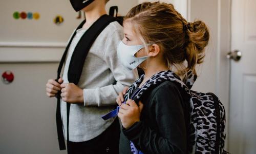 Two children stand side by side wearing backpacks and masks.