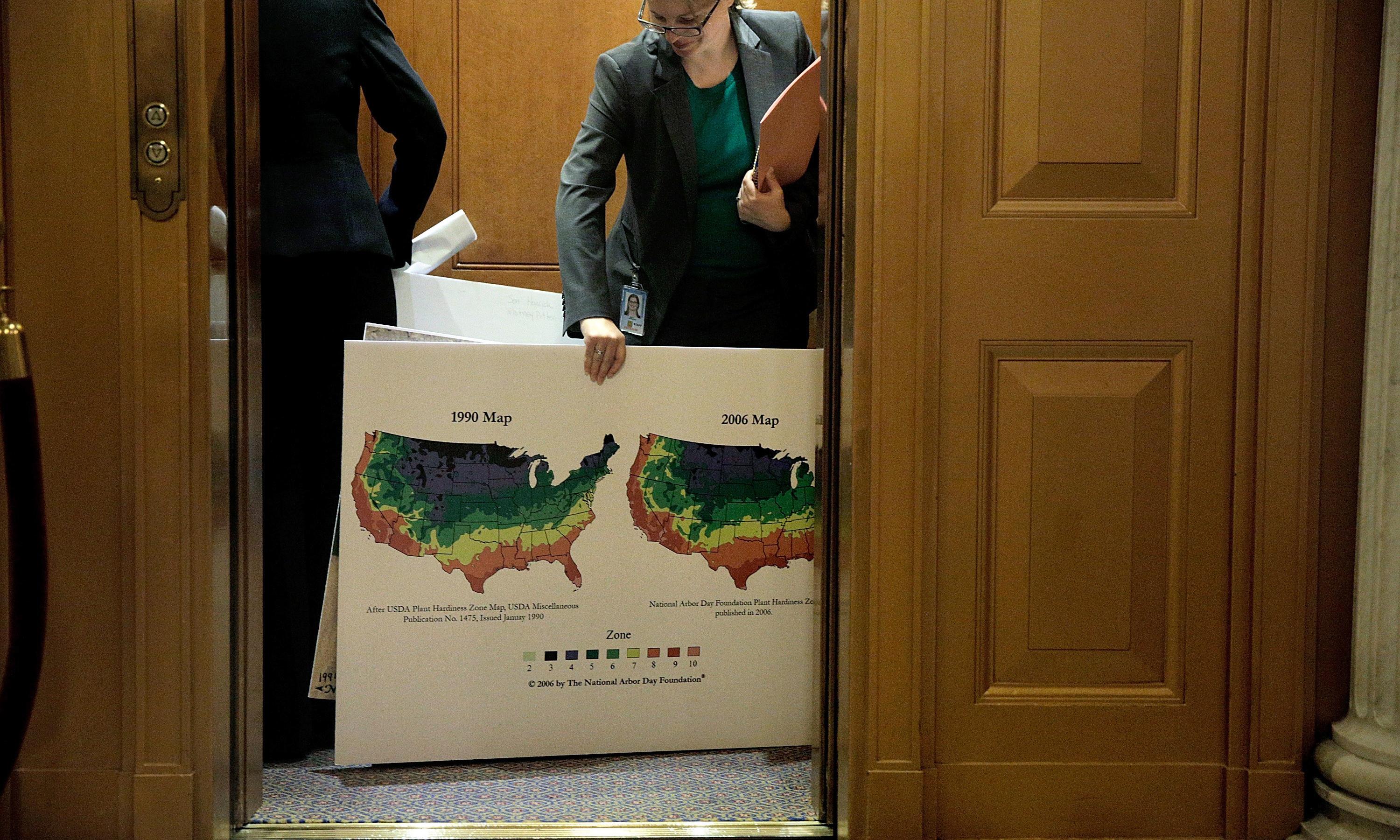 A woman entering a hearing holding a sign that shows two maps that compare plant hardiness zones.