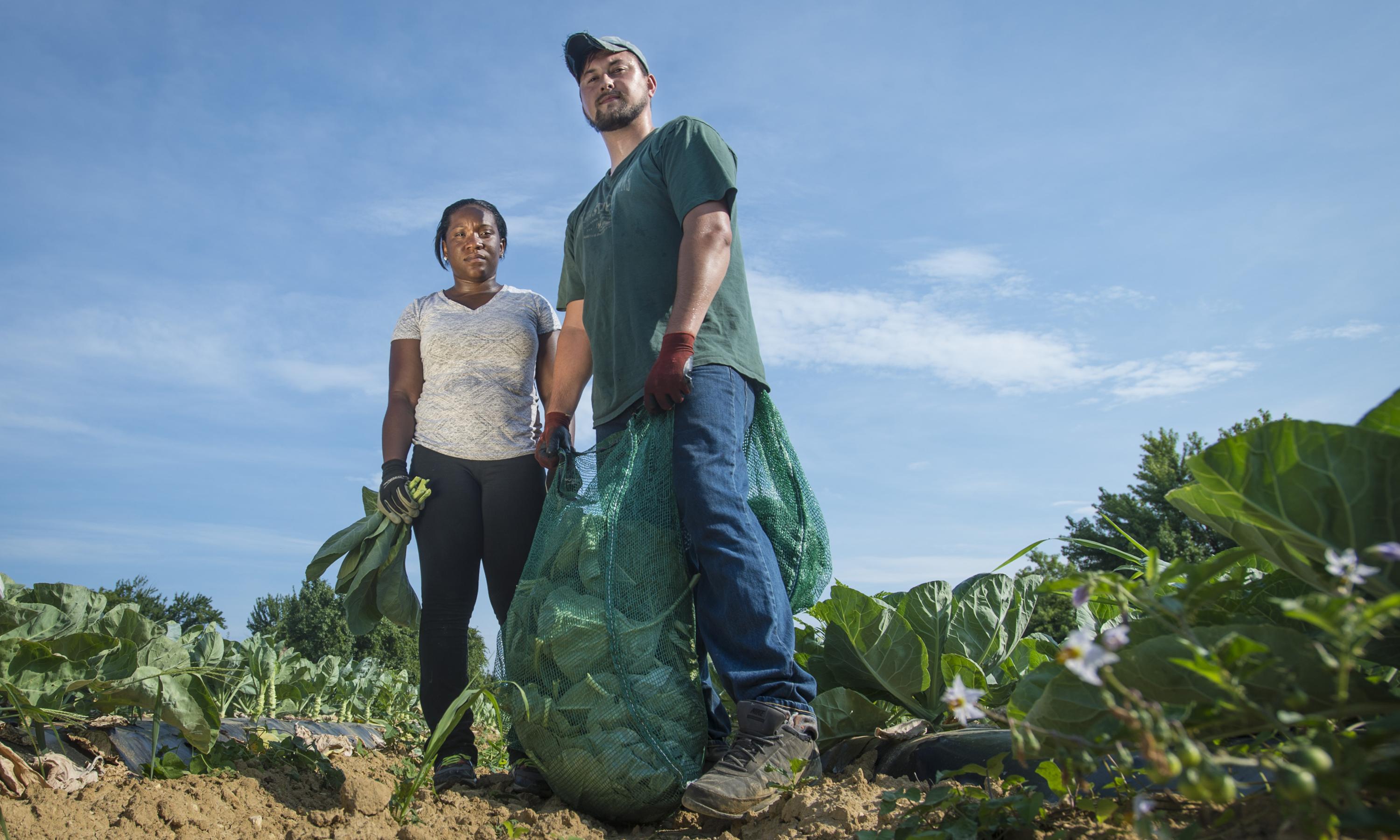 Two farmers stand side by side, both holding crops